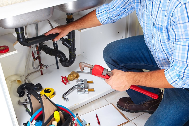 Plumber on the kitchen. Why You Should Never Ignore Slow-Draining Kitchen Plumbing. Photo of a plumber performing maintenance under a kitchen sink.